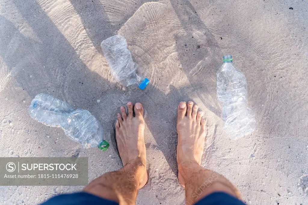 Man's feet on the beach surrounded by empty plastic bottles, top view