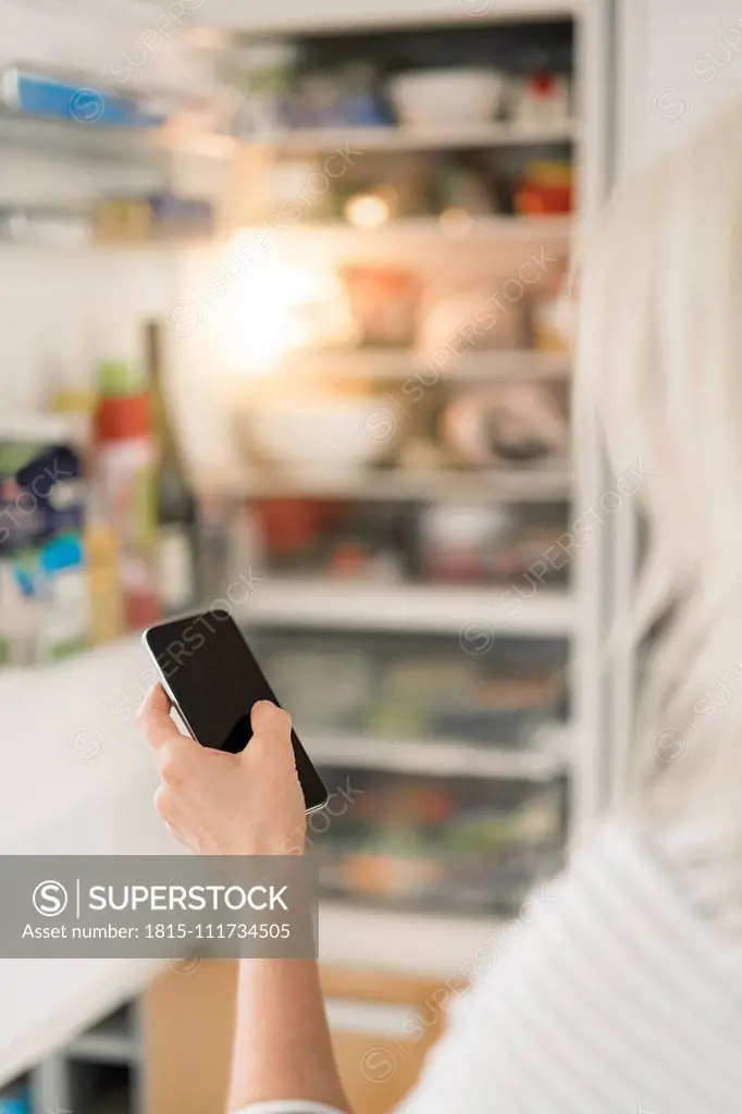 Hand of woman with smartphone checking fridge in kitchen at smart home