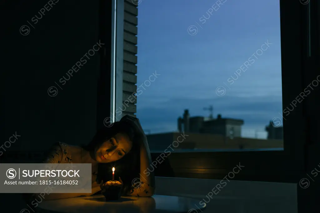 Lonely birthday woman looking at burning candle on cupcake by window in room during sunset
