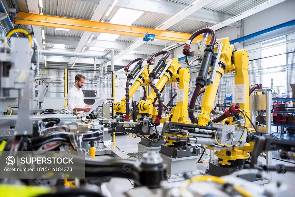 Young engineer reading paper while standing by robotic arms in automated industry