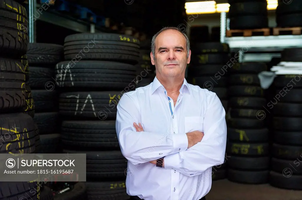 Portrait of confident senior male owner standing with arms crossed against tire at store