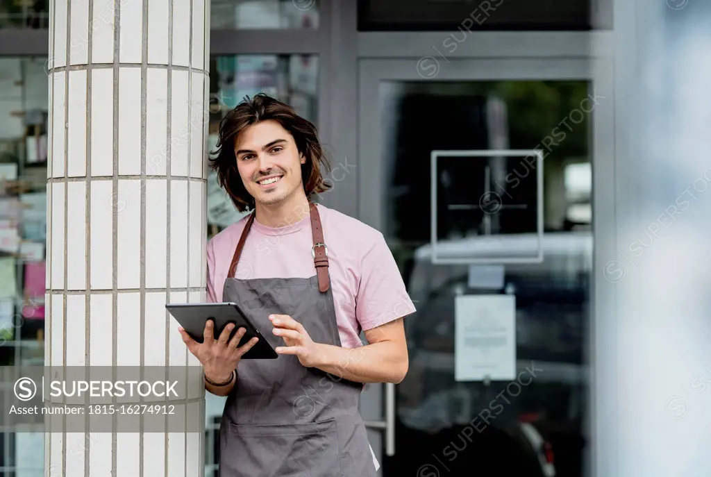Smiling owner using digital tablet while standing by column against cafe