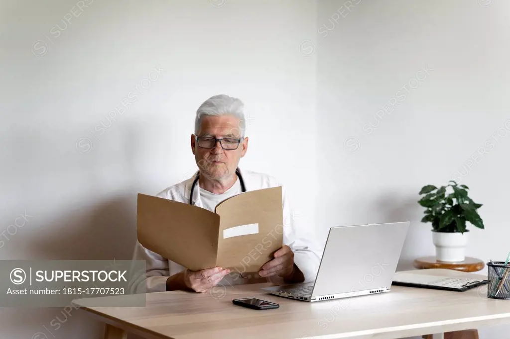 Senior doctor examining medical record while sitting at desk in clinic