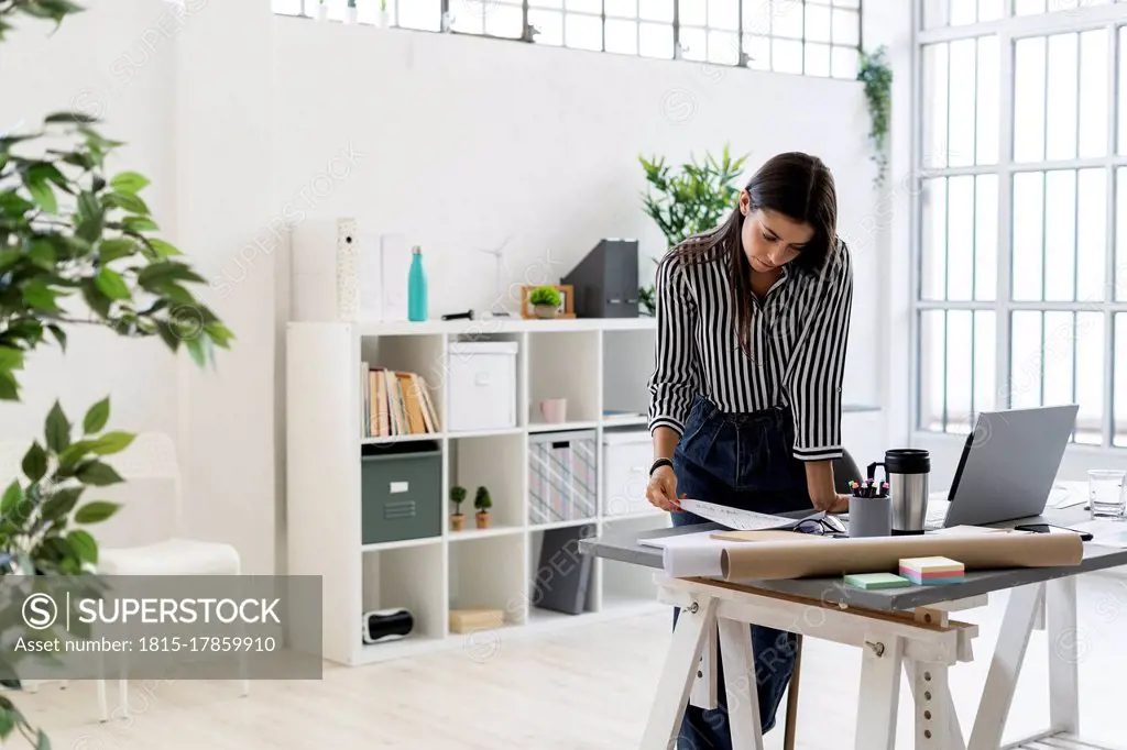 Young female design professional examining plan at desk while working in creative office