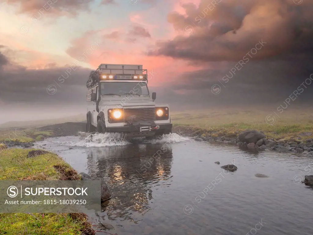 Off-road vehicle splashing water from puddle against cloudy sky at sunset