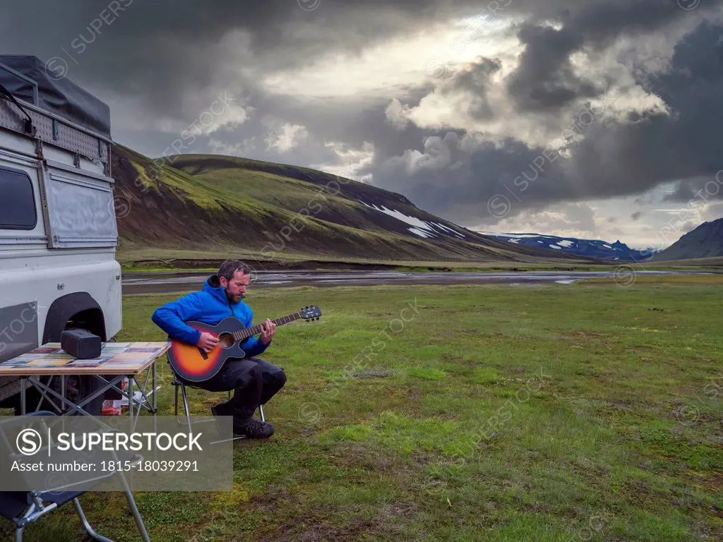 Man playing guitar while relaxing by off-road vehicle against cloudy sky during sunset
