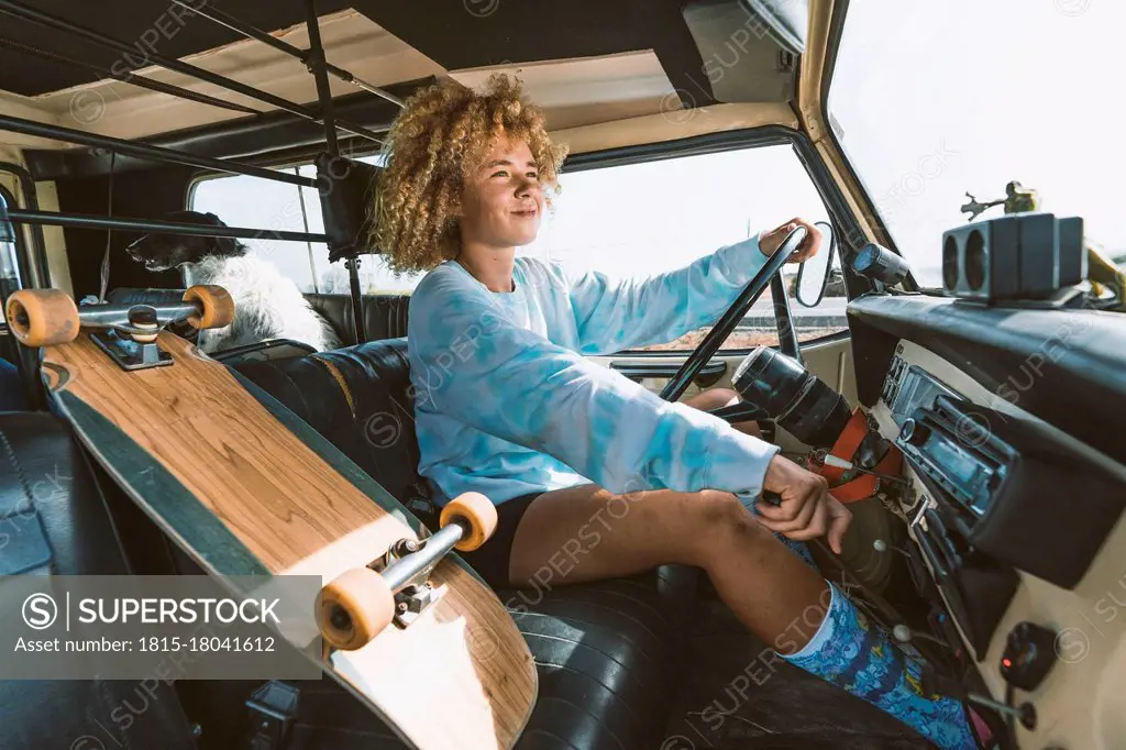 Smiling blond Afro woman sitting by skateboard while driving old off-road vehicle