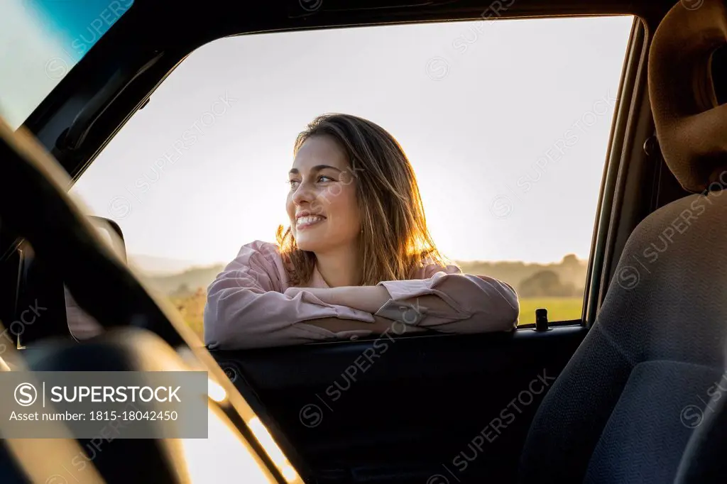 Happy young woman looking away while leaning on car window during sunset