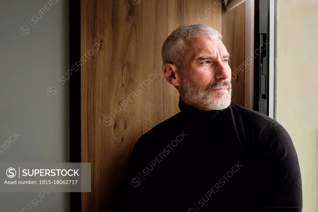 Mature man in warm clothing day dreaming while looking through window in living room