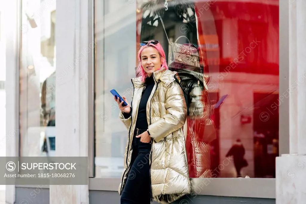 Fashionable woman using mobile phone while standing with hands in pockets against window