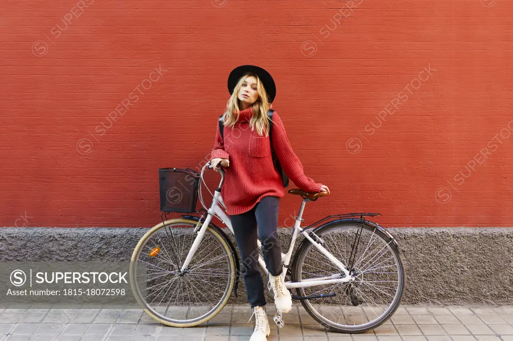 Young blond woman wearing hat standing with bicycle against red wall