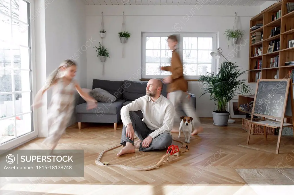Siblings running around father sitting on floor in living room at home