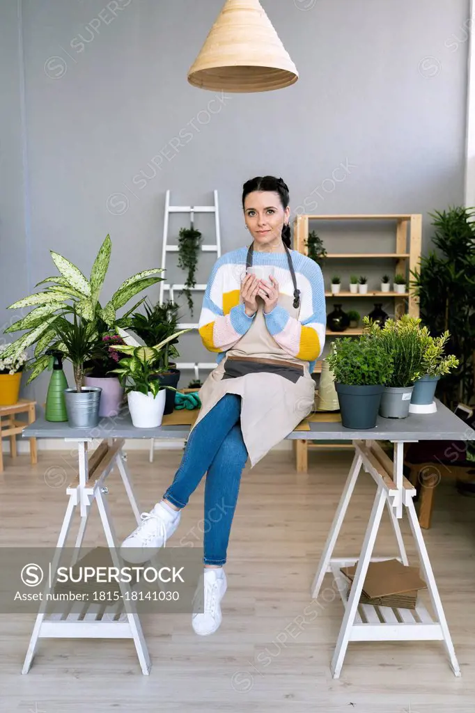 Young woman holding mug while sitting on table in workshop