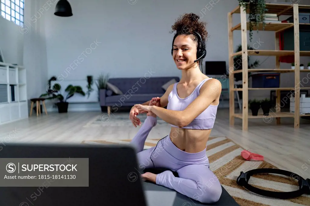 Smiling yoga influencer with headphones doing mermaid pose while vlogging through laptop at home