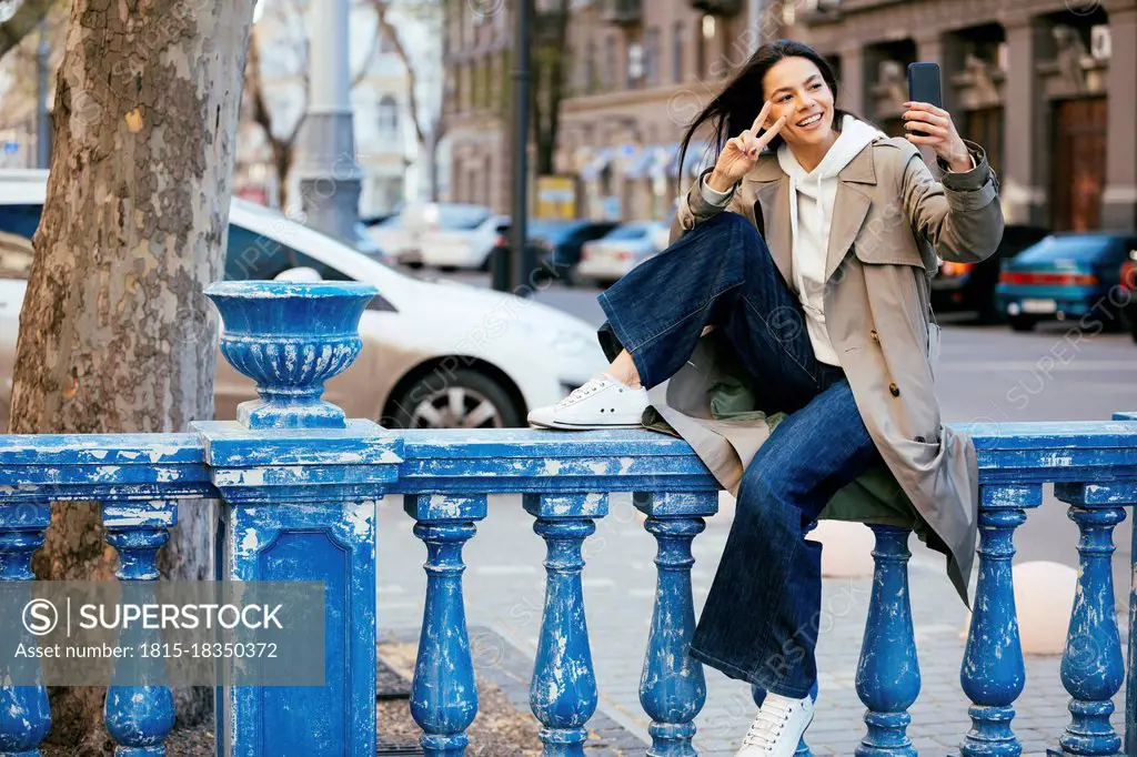 Smiling woman sitting on blue railing showing peace sign while taking selfie