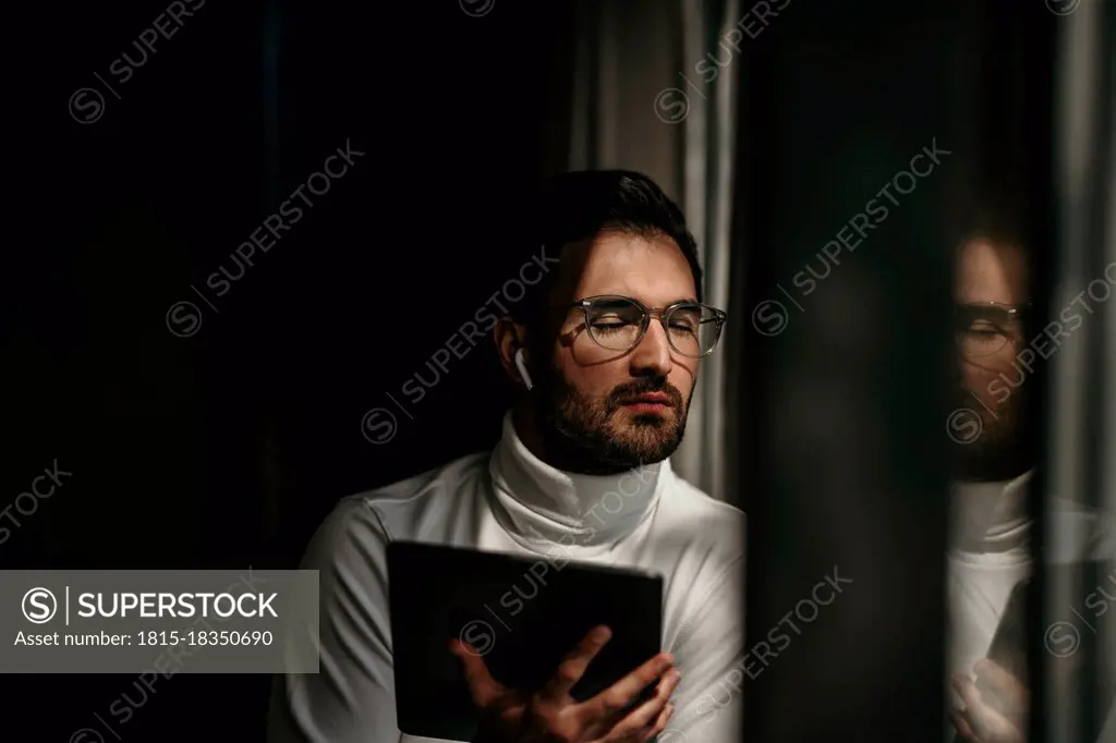 Businessman with eyes closed holding digital tablet by window at home