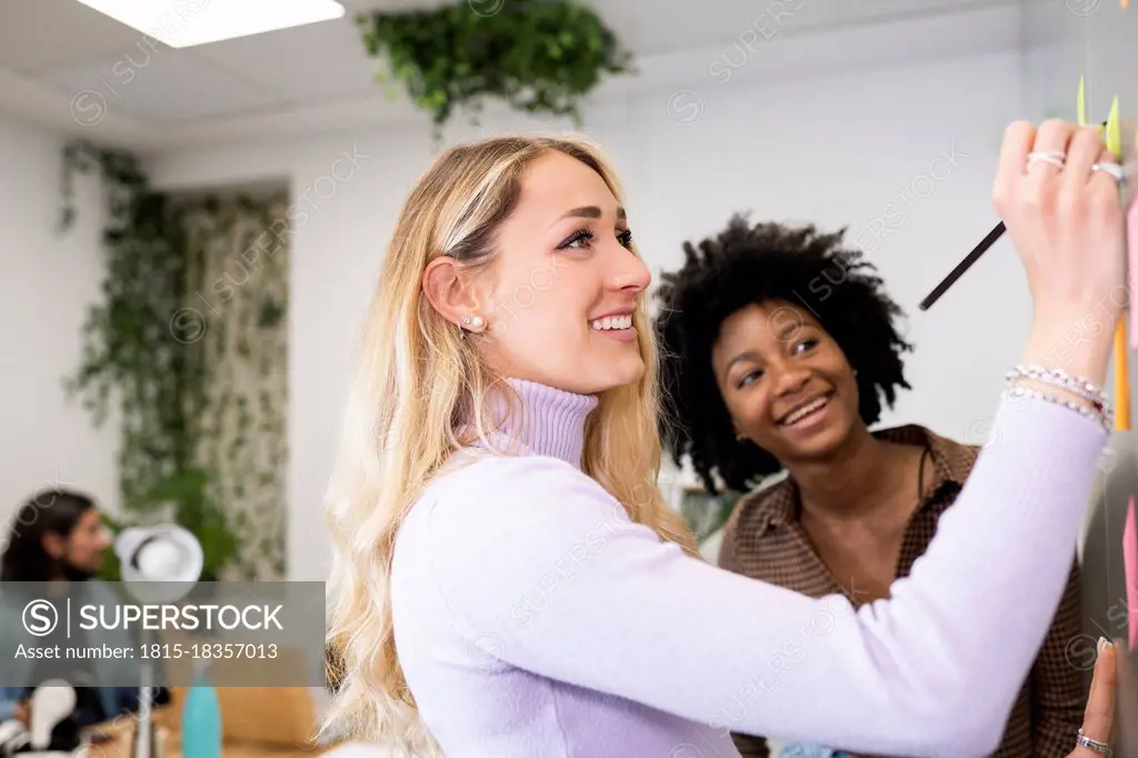 Smiling young businesswoman writing on adhesive notes while discussing with female coworker in office