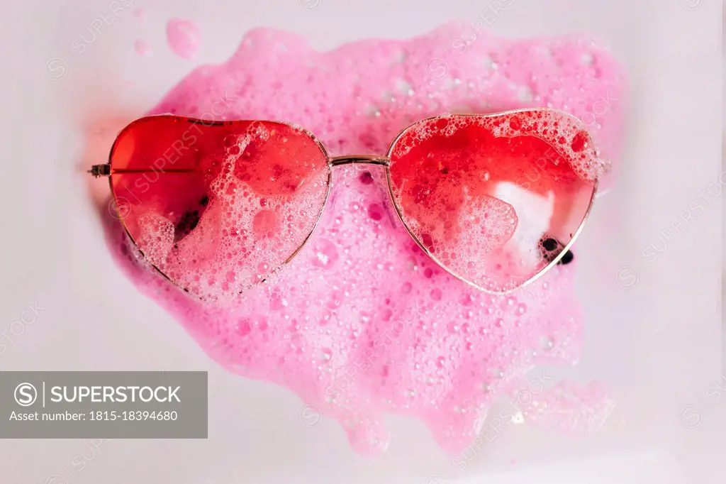 Red heart shape sunglasses on pink soap sud over white background