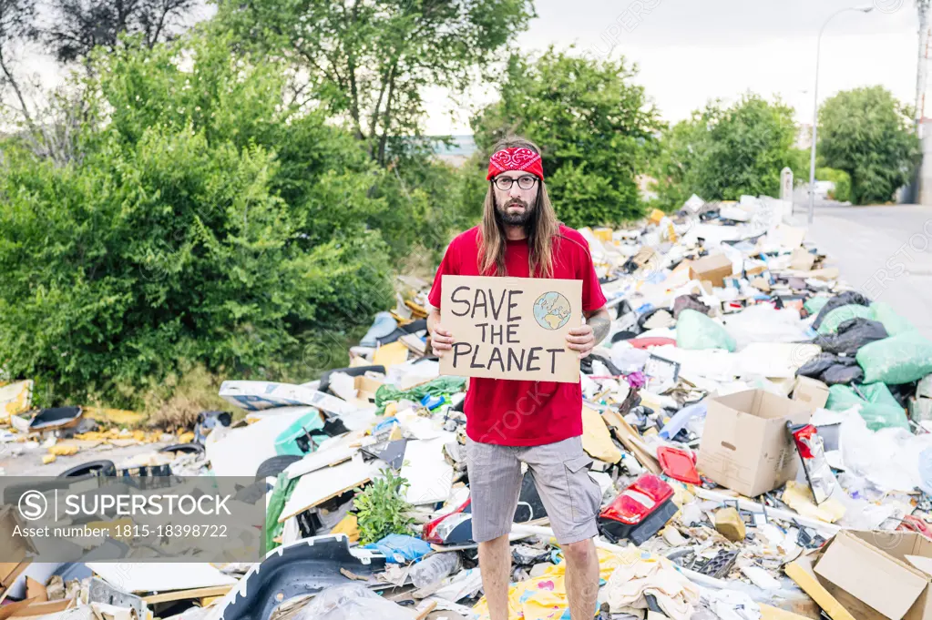 Young hippie man holding save the planet cardboard while standing on garbage dump