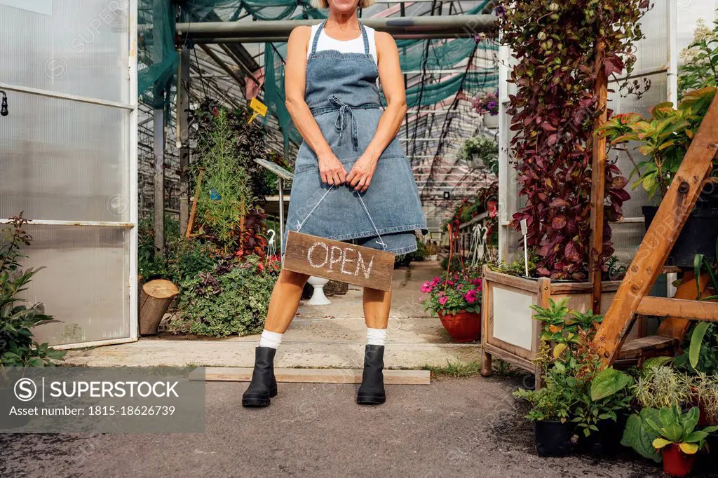 Female greenhouse worker holding wooden open sign at doorway of greenhouse