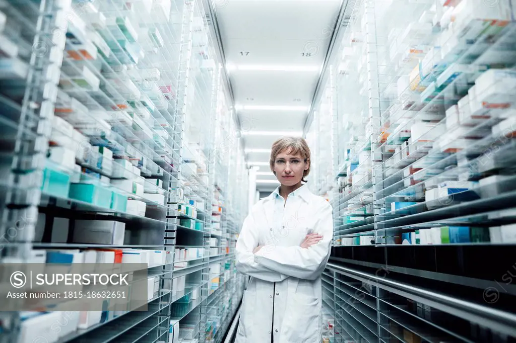 Confident female pharmacist with arms crossed standing amidst racks at medical store