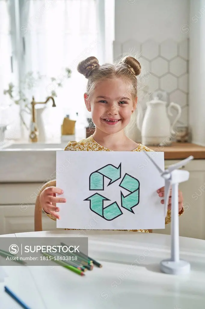 Smiling girl holding recycling symbol drawing on paper at table