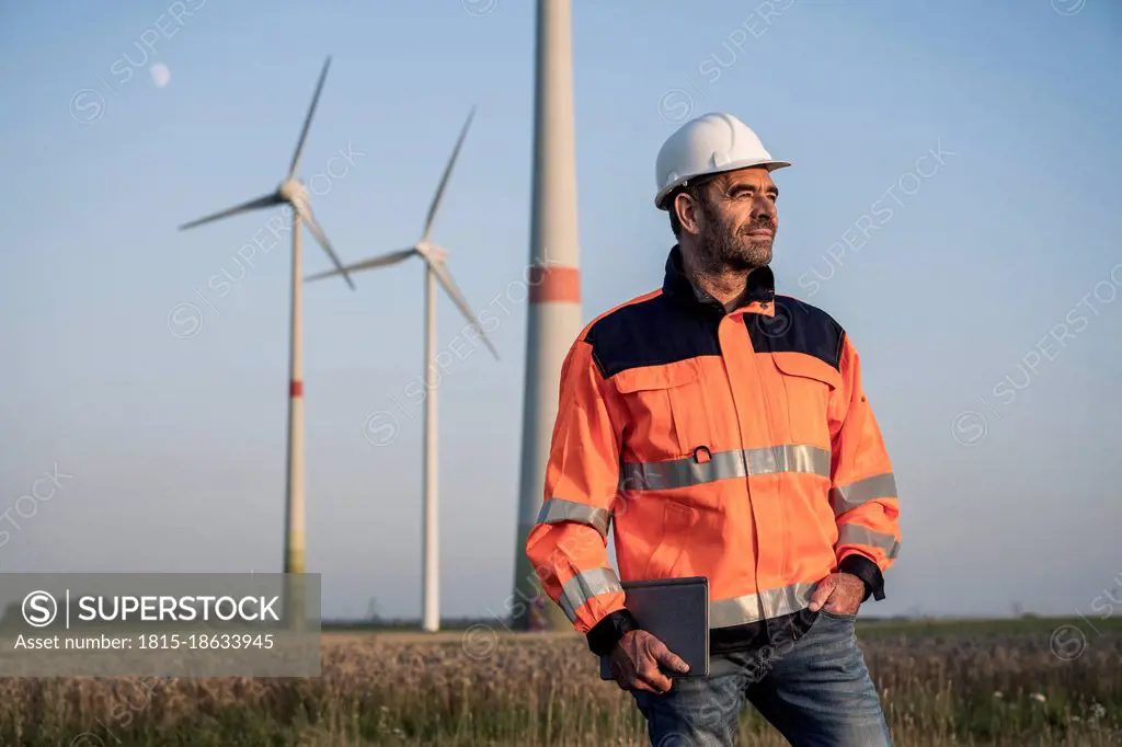Male engineer holding digital tablet while standing in front of wind turbines
