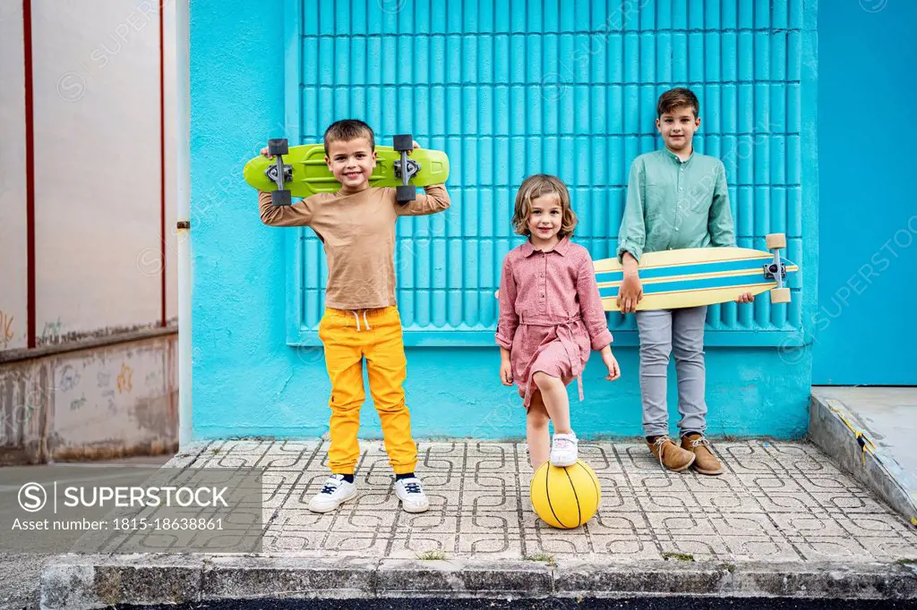 Children standing with skateboards and sports ball on footpath