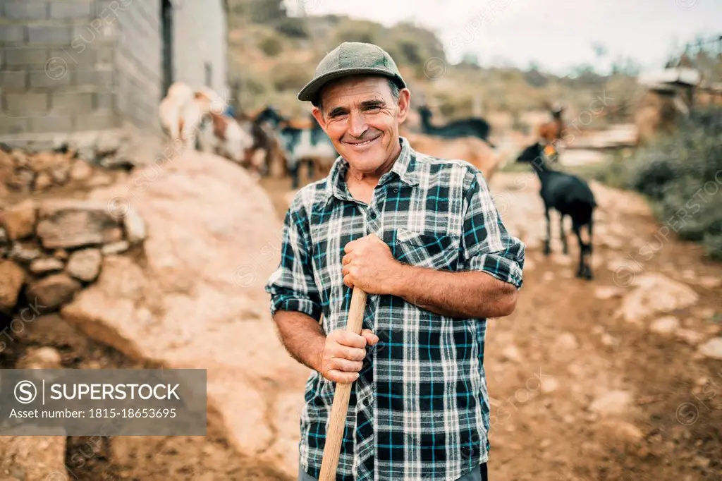 Smiling male goat herder holding broom while standing at farm