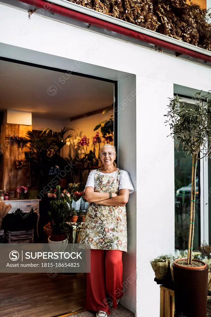 Smiling female florist standing with arms at flower shop entrance