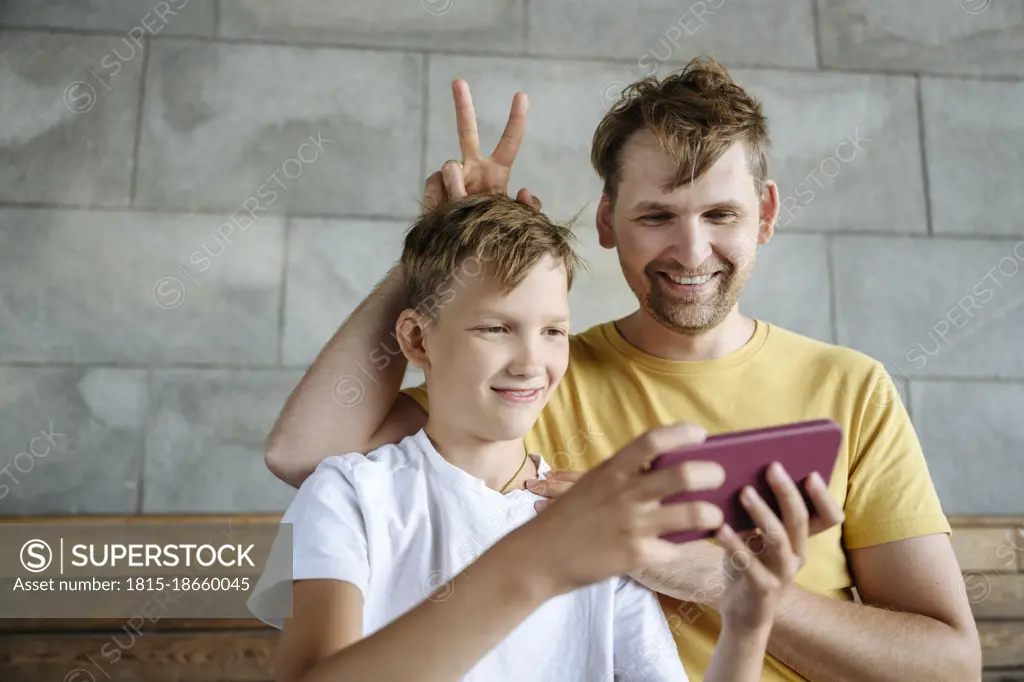 Smiling man gesturing while son taking selfie through mobile phone on bench