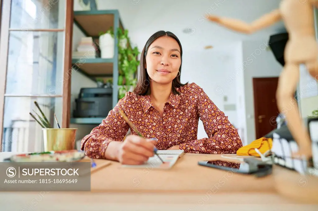 Young female cartoonist holding paintbrush while sitting on desk