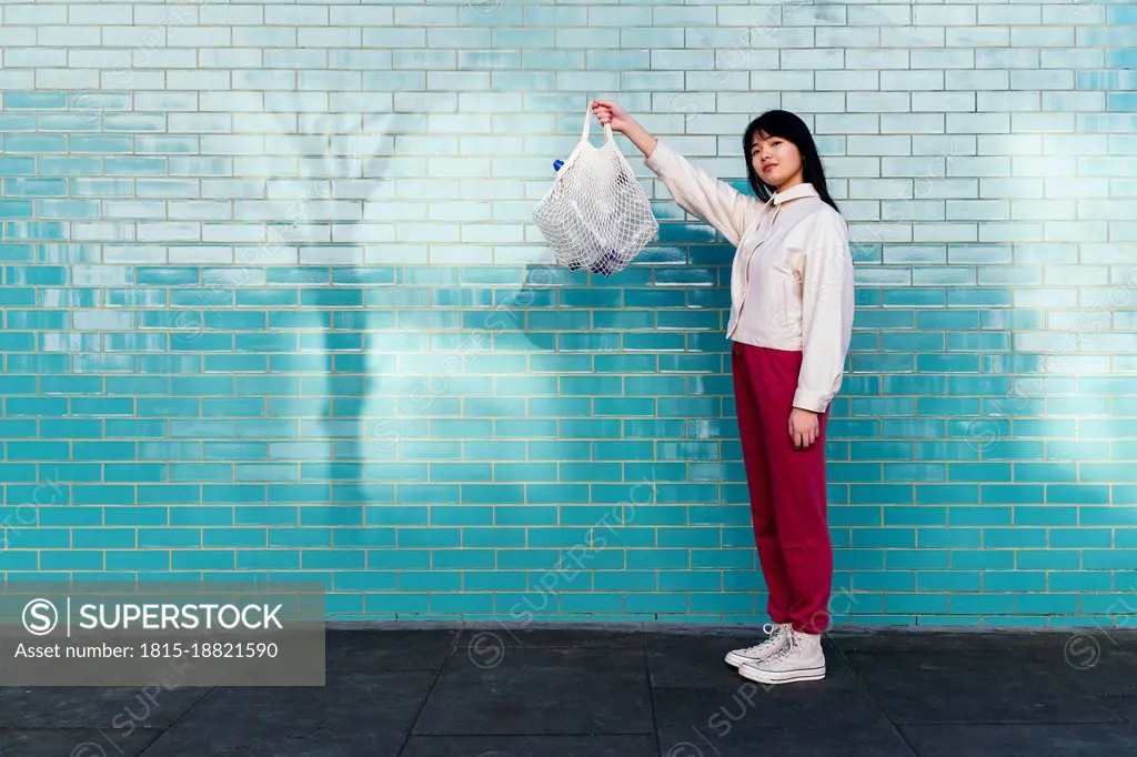 Young woman showing mesh bag with empty plastic bottles standing by turquoise brick wall