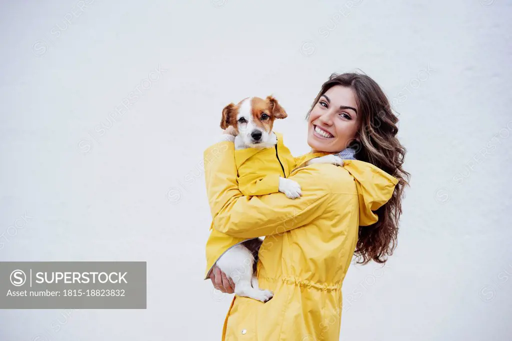 Smiling young woman embracing dog by white wall