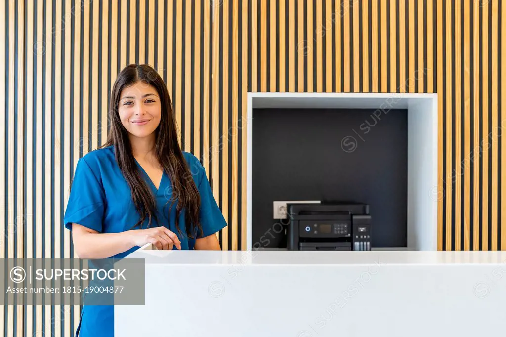 Smiling nurse with long hair standing by reception desk at hospital
