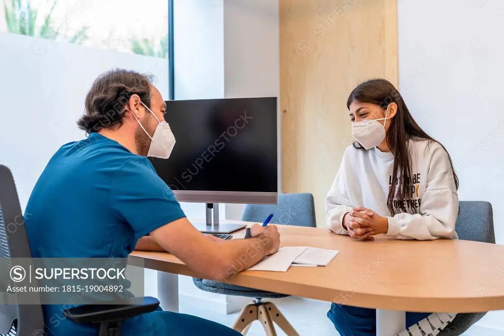 Doctor discussing with patient sitting at desk in hospital during pandemic
