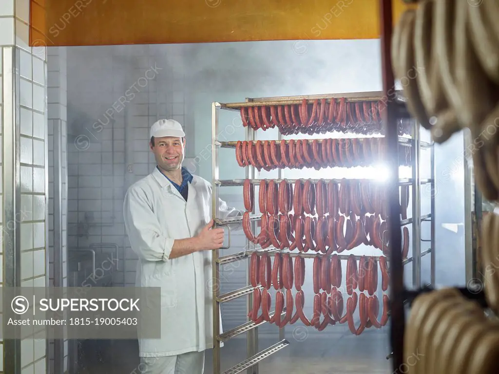 Smiling butcher standing by arranged sausages in smokehouse