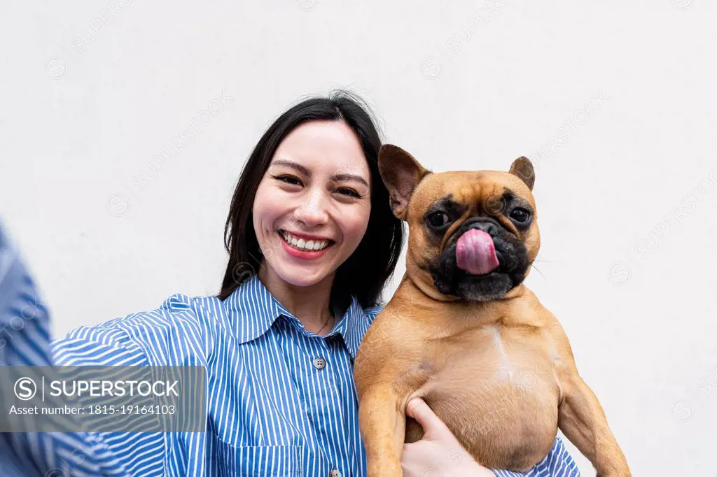 Happy woman carrying dog sticking out tongue in front of wall