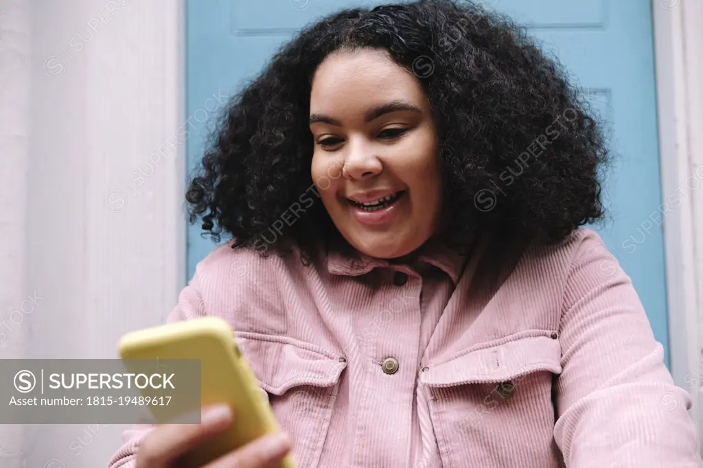 Happy young woman with curly hair surfing net through smart phone