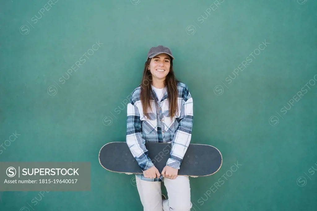 Smiling woman with skateboard standing in front of green wall