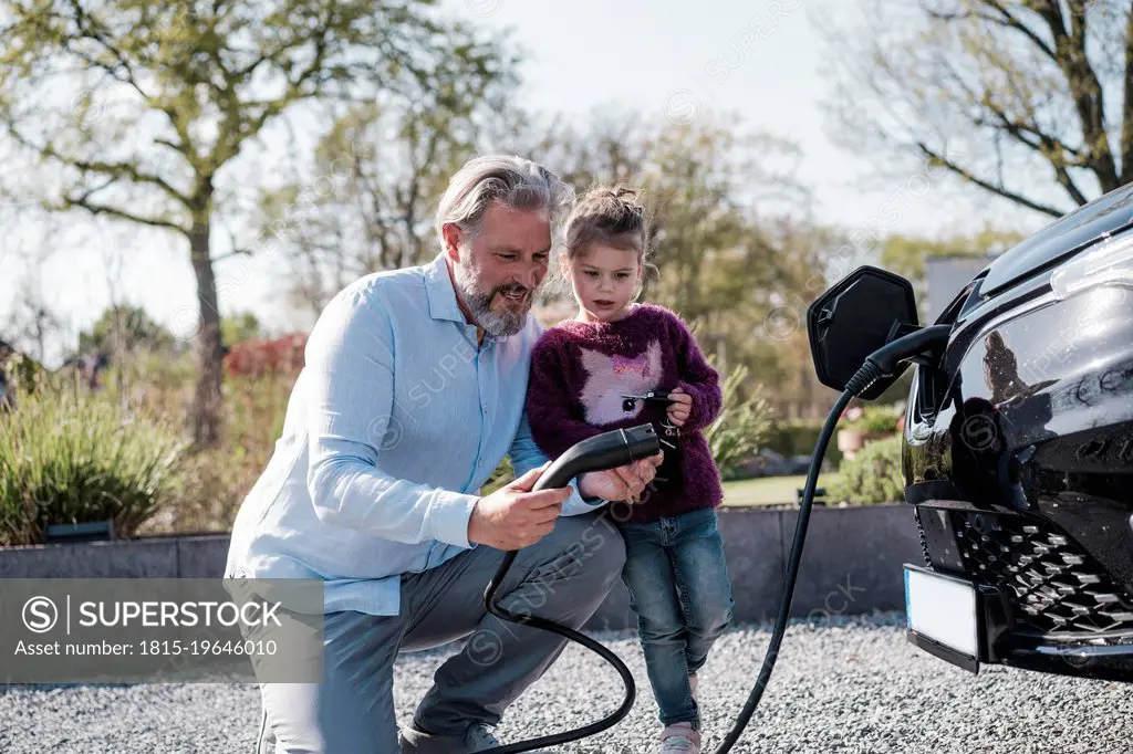 Man showing electric plug to daughter standing in front of car on sunny day