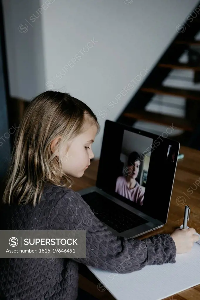 Girl writing on paper studying through video call on laptop at home