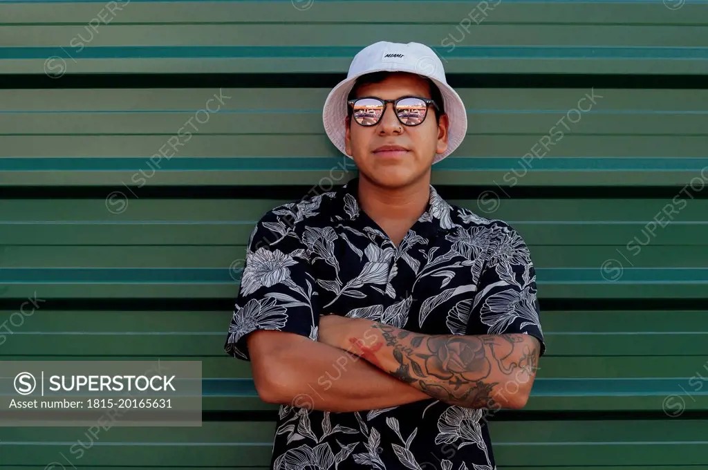Young man wearing bucket hat standing with arms crossed in front of green wall