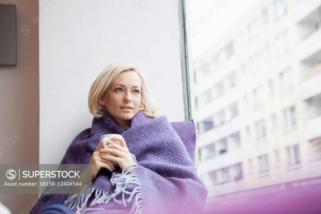 Portrait of woman with cup and woolen blanket looking through window