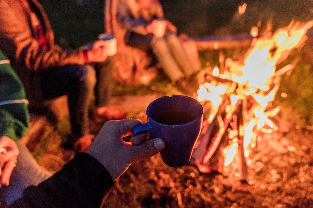 Hand of person holding tea cup, group of people sitting at a camp fire