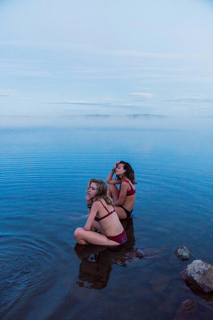 Two young women wearing bikinis, sitting on a stone in a lake