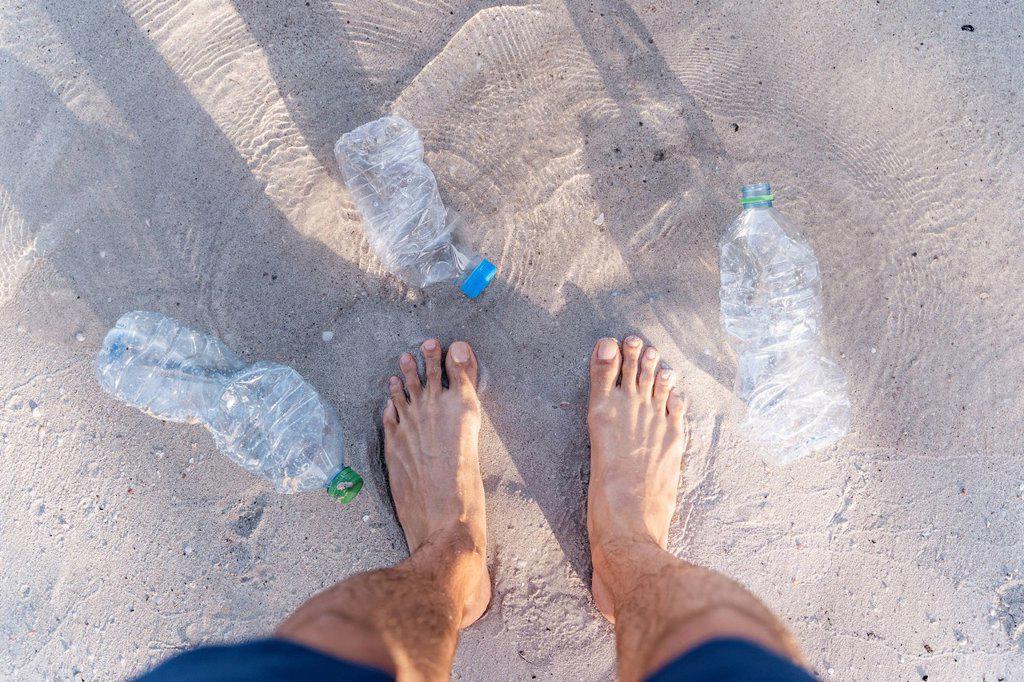 Man's feet on the beach surrounded by empty plastic bottles, top view
