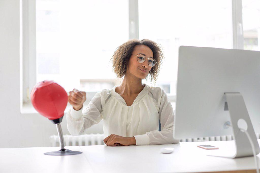 Portrait of young freelancer sitting at desk in office boxing punching ball while looking at computer