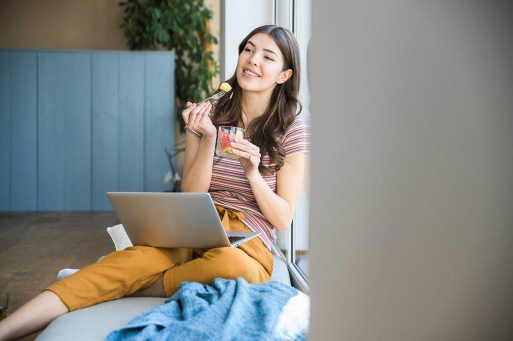 Young woman sitting at the window at home with laptop having a snack
