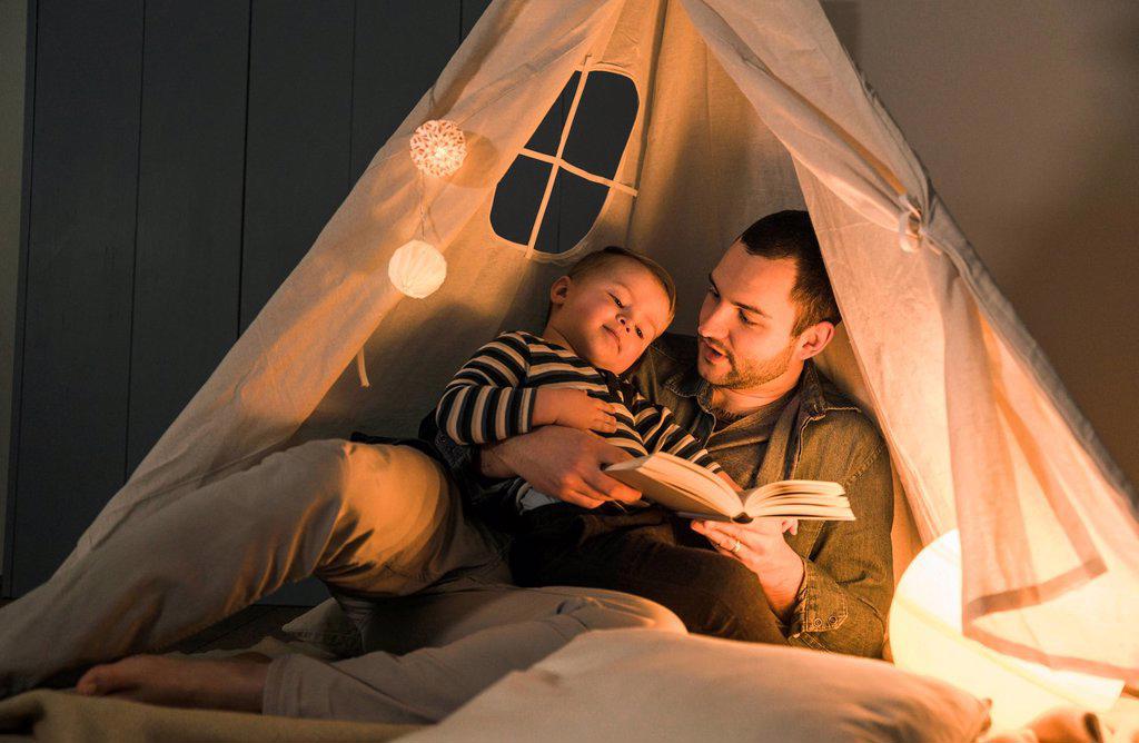 Father reading book to son at an illuminated tent at home